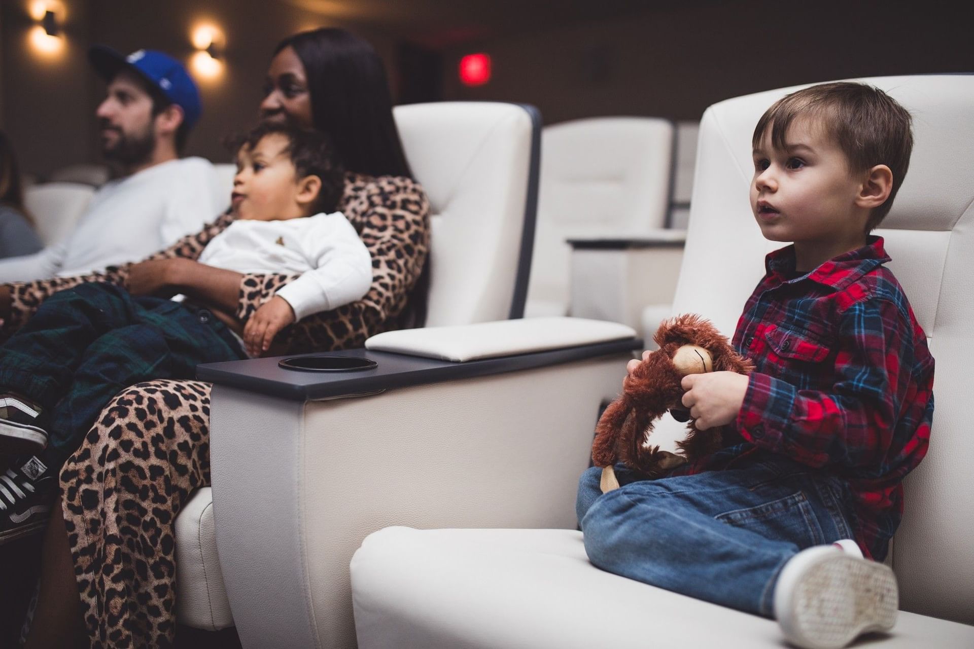 Young boy holding a plush toy monkey sits on white cinema seats with his family in a screening room at Hotel X Toronto