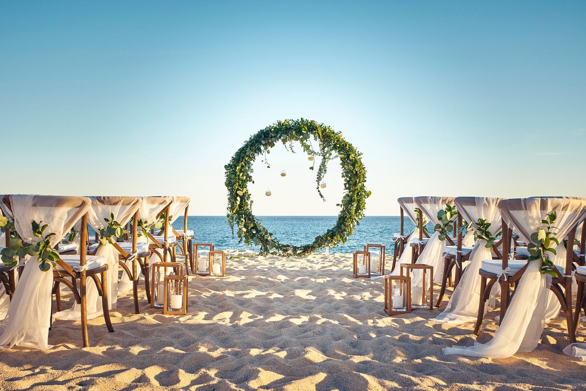 Elegant beachfront wedding with sheer-wrapped chairs facing a floral arch on the sand at Hacienda Del Mar Los Cabos