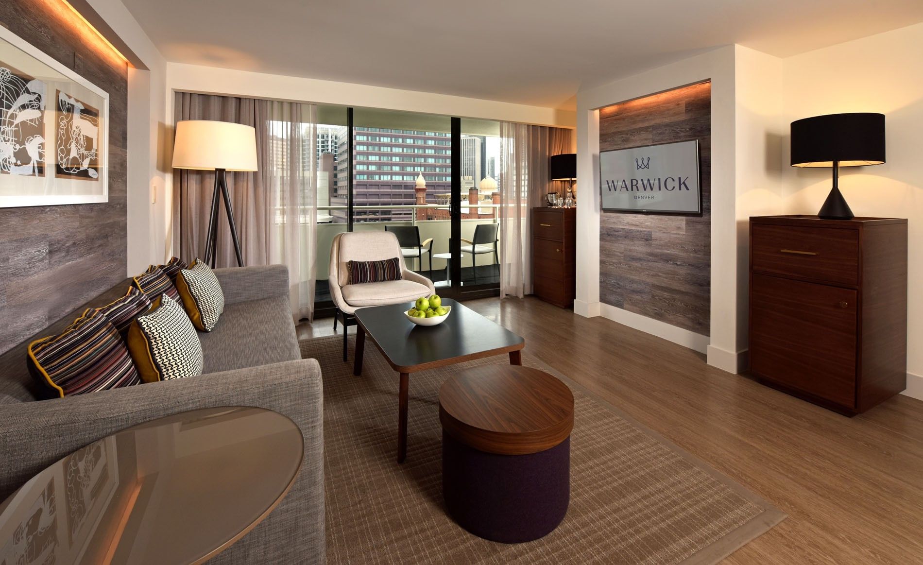 A wide view of a hotel suite with a sofa, coffee table, armchair, and balcony with a city view.