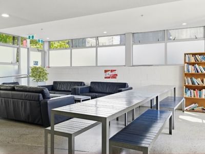 Modern lounge area with black couches, a long table, and a bookshelf at Student Living Auckland – Anzac.