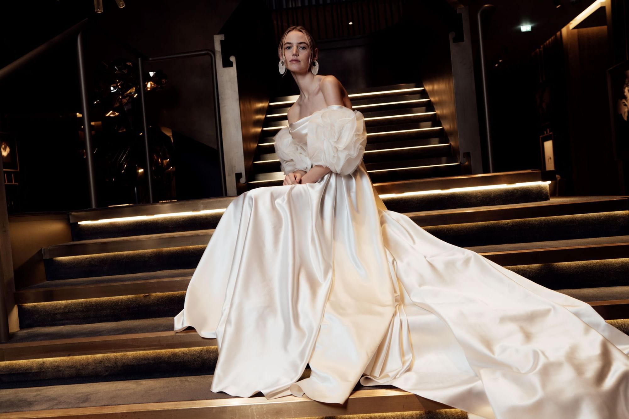A lady with a white wedding dress posing for a picture on stairs in a dark background at The Londoner Hotel