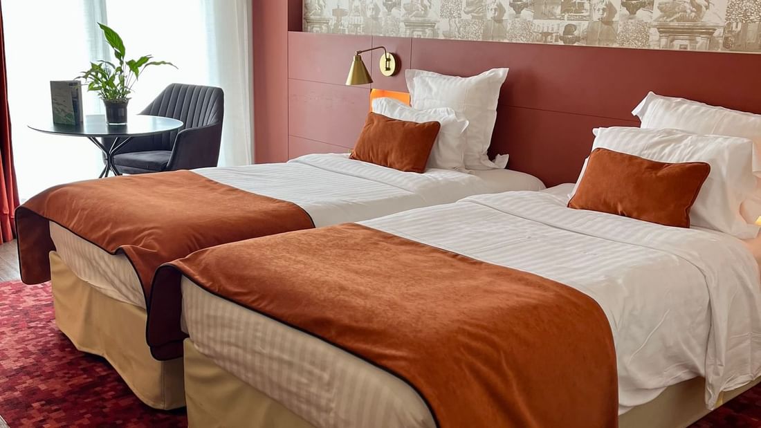 Two beds with orange blankets and white sheets on a red rug in a hotel room.