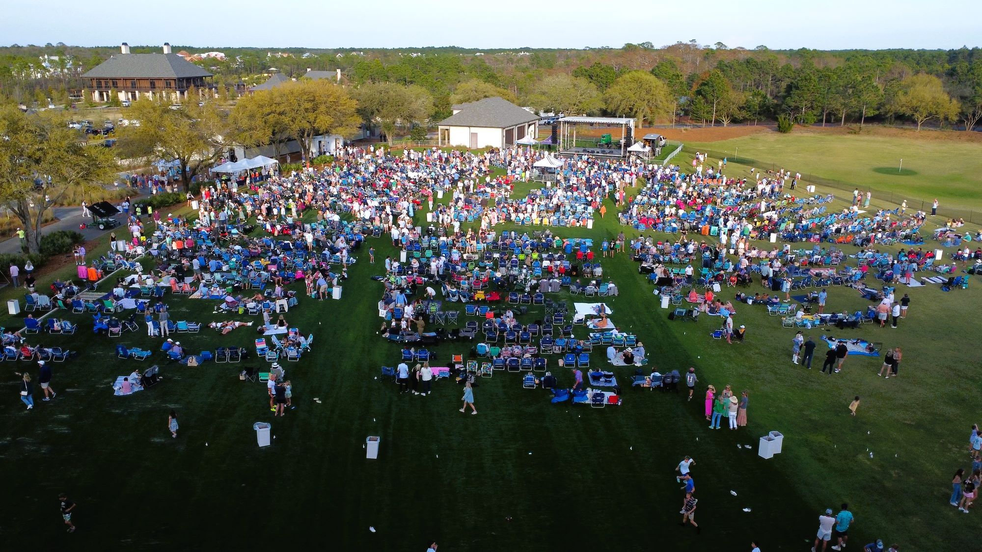Aerial view of a large crowd seated on grass with a stage in the background.