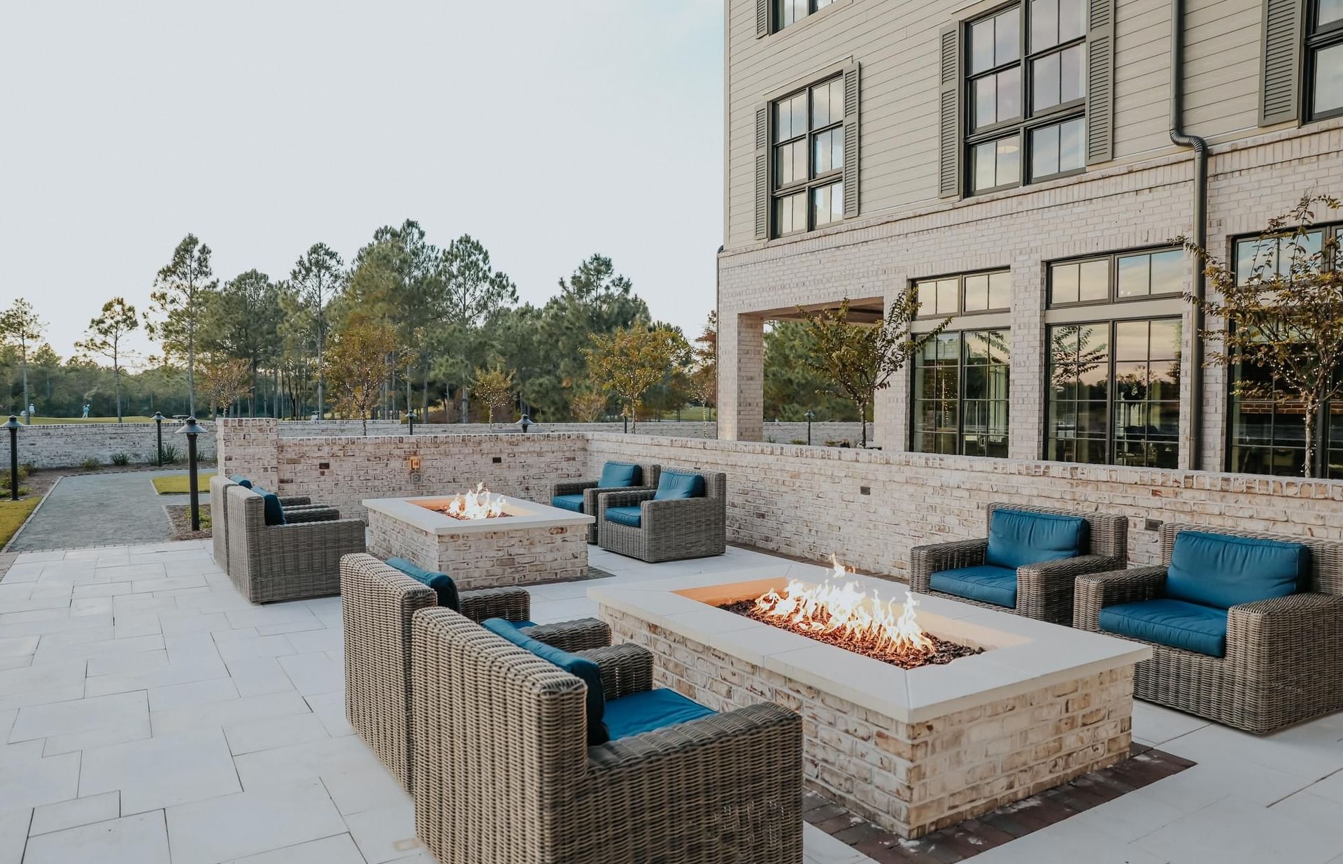 Outdoor patio with wicker chairs, blue cushions, and two fire pits surrounded by brick walls and building.