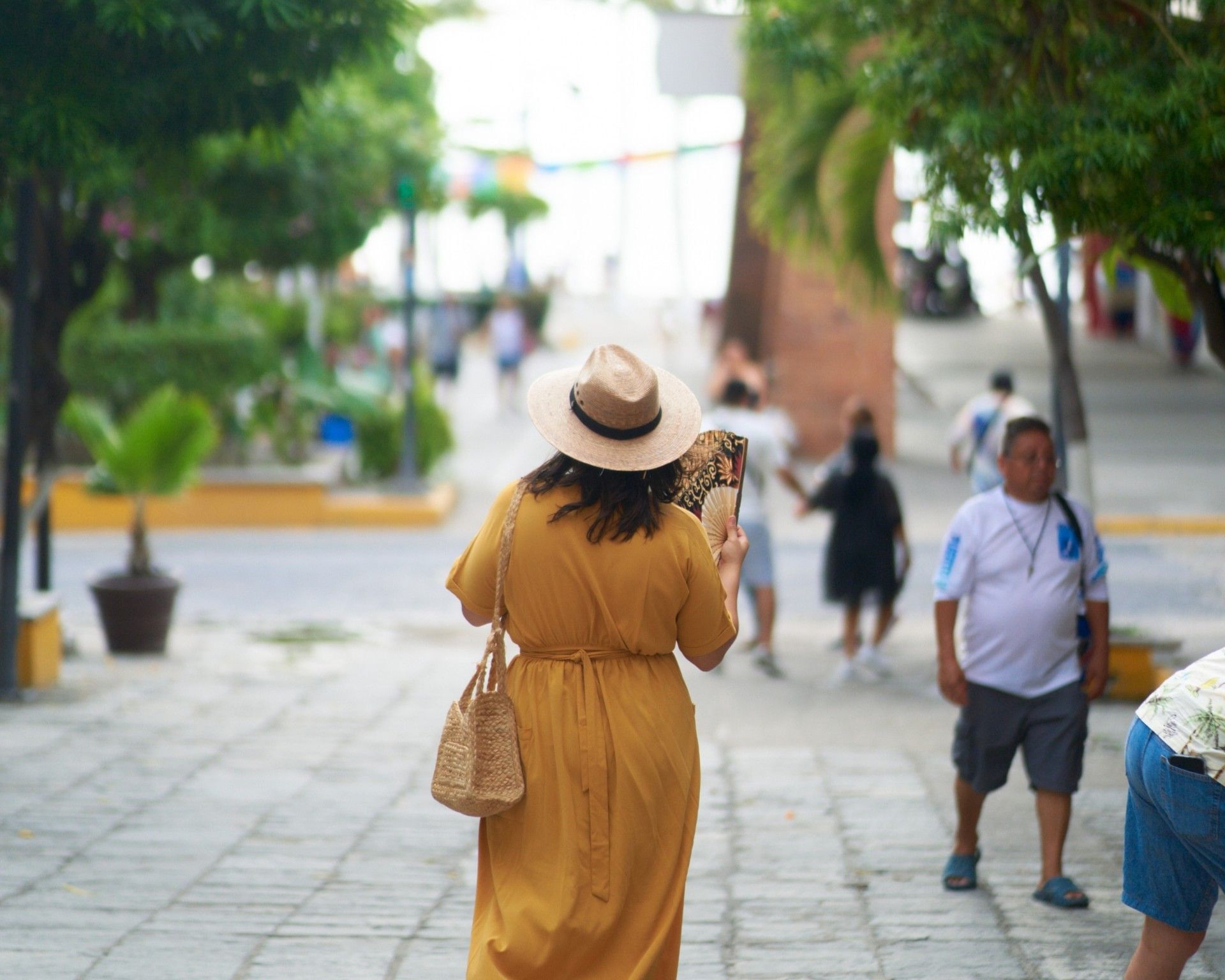 Una mujer con vestido amarillo mostaza, sombrero de paja y bolso tejido camina por una calle peatonal empedrada en Puerto Vallarta, México, sosteniendo un abanico decorativo mientras otras personas pasean entre árboles frondosos y edificios coloridos al fondo.