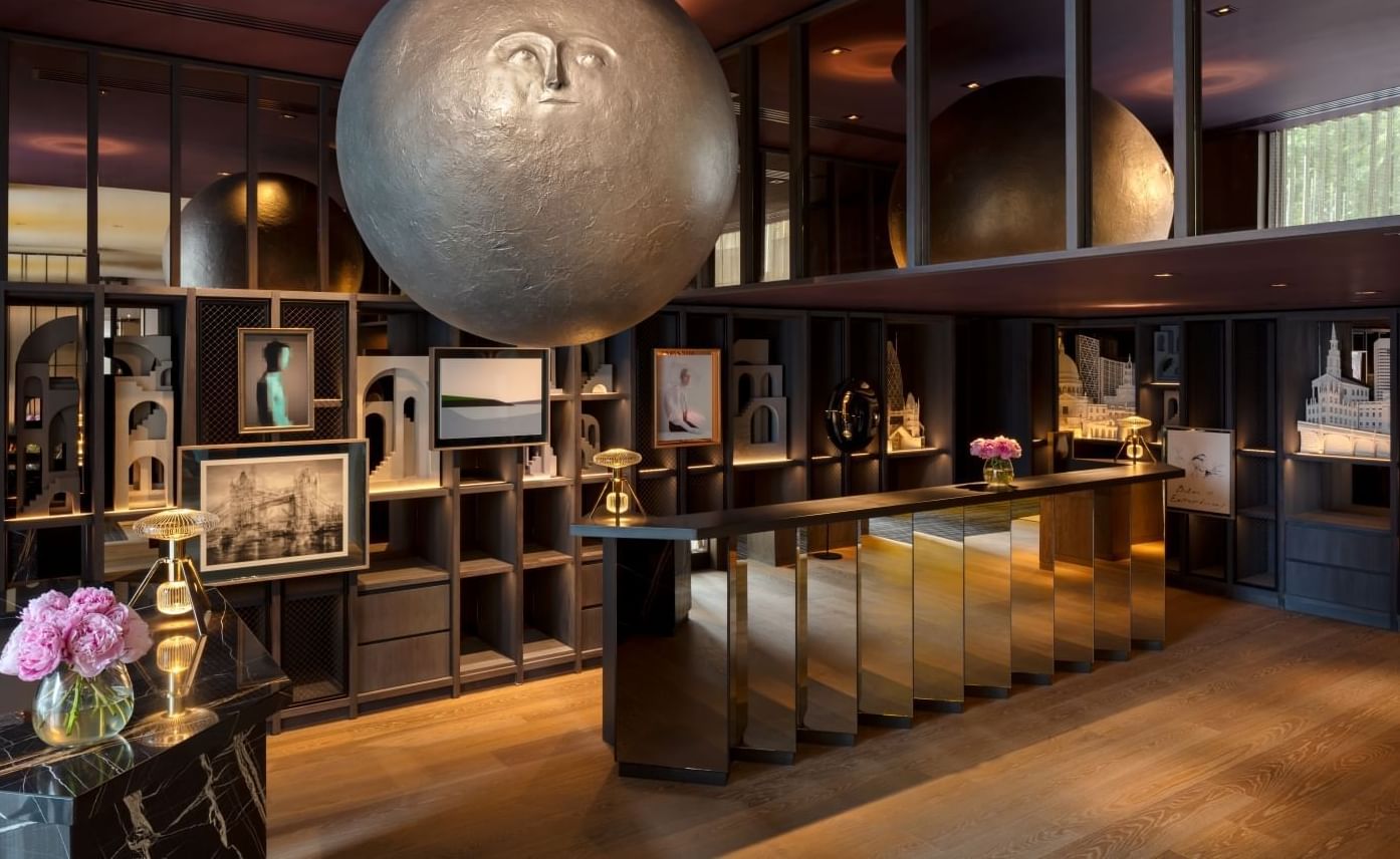 Interior luxuoso do átrio do The Londoner, com uma grande escultura esférica, balcão de receção espelhado e detalhes florais