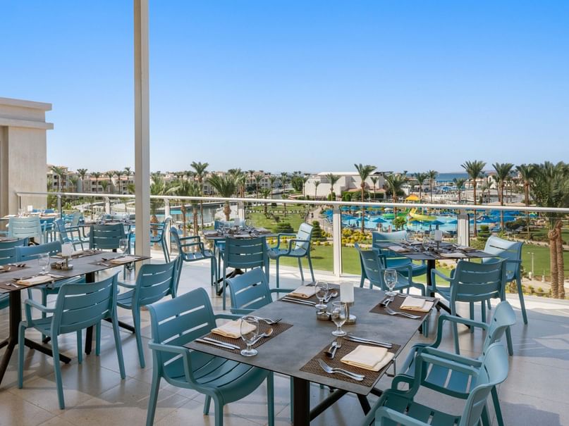Outdoor dining area with blue chairs and set tables, overlooking a resort with pools and palm trees.