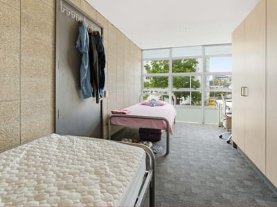 Two beds with white mattresses and pink sheets in a room with clothes hanging and large windows.