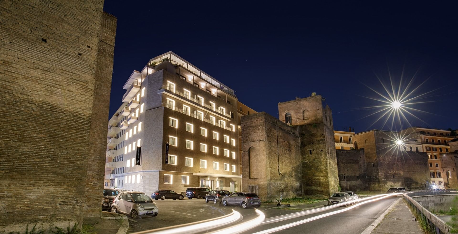 Tribune Hotel Rome exterior at night with modern illuminated façade beside ancient Roman walls, blending contemporary design with historic surroundings.