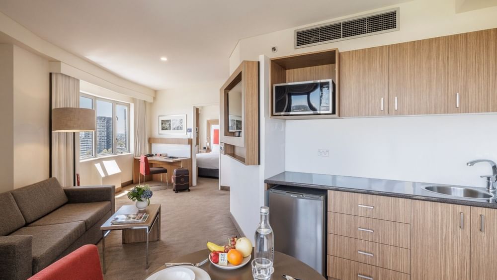 Modern suite with kitchenette, living area, and bedroom visible through doorway with city views from large windows