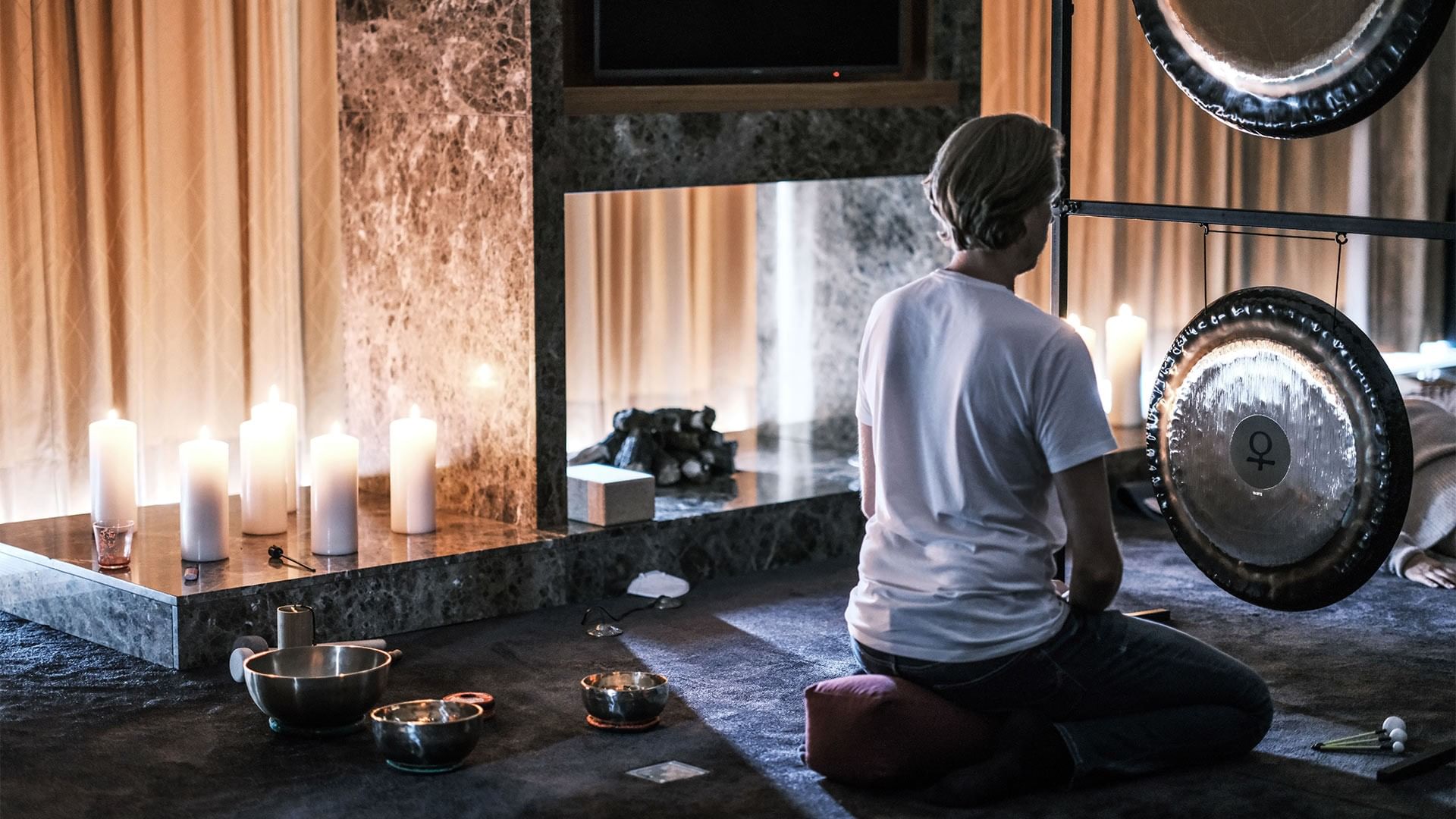 Person meditiert vor Gongs, Kerzen und einem Kamin in einem Wellnessbereich.