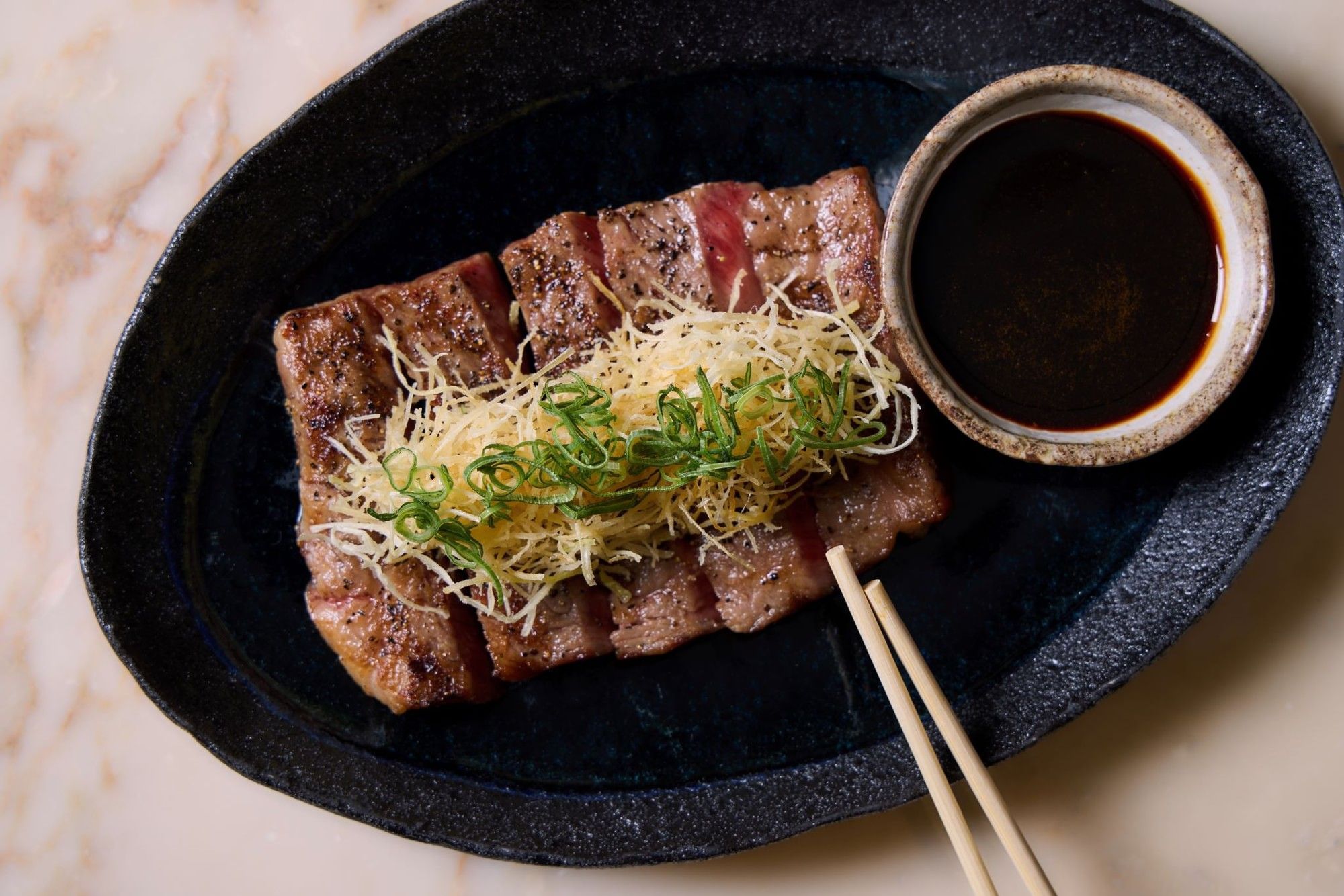 Sliced ribeye steak with toppings and dipping sauce on a plate.