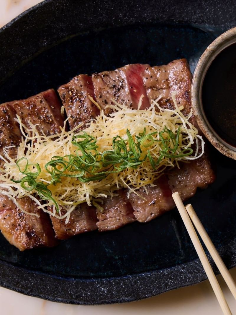 Sliced ribeye steak with toppings and dipping sauce on a plate.