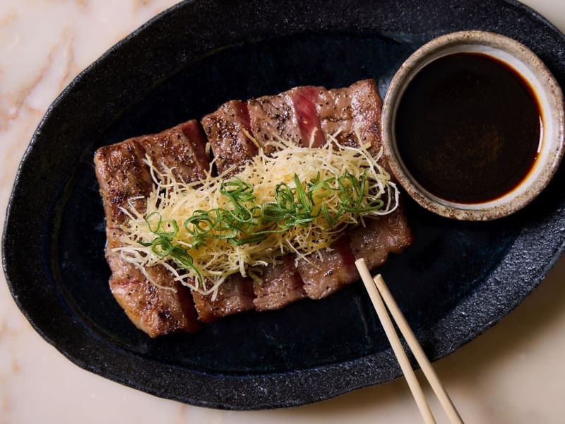 Sliced ribeye steak with toppings and dipping sauce on a plate.