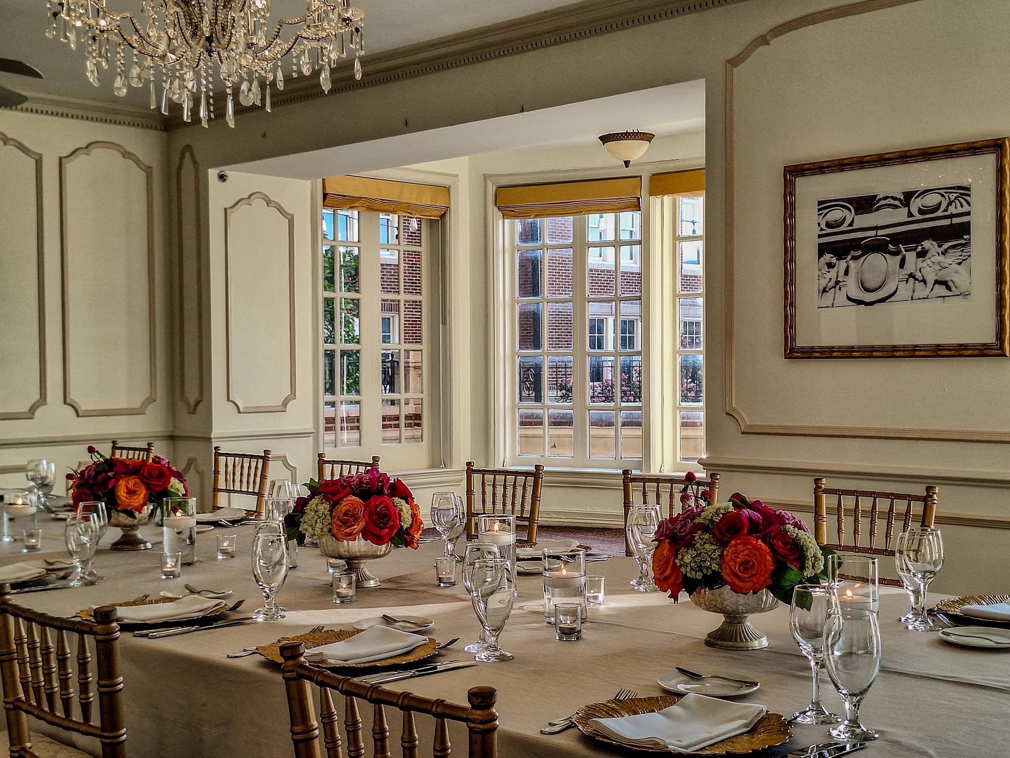 A formal dining room with a long table set for a meal, featuring a chandelier, floral arrangements, and large windows.