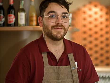 Barman tatoué avec tablier de cuisine et lunettes devant des étagères de bouteilles à l'Hôtel Rivella.