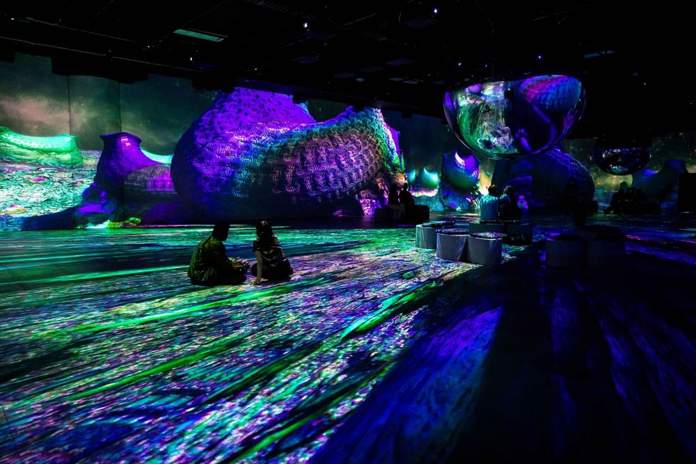 Two visitors sit on the floor viewing an immersive digital art projection with vibrant purple and green lighting at Warwick Le Crystal Montreal