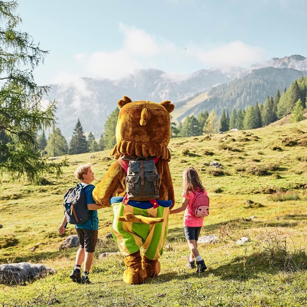 Two children walking with a mascot on a mountain trail.