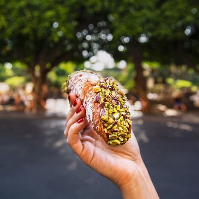 Eine Hand hält ein Gebäck mit Pistazien und Zucker, vor Bäumen und einer Straße.