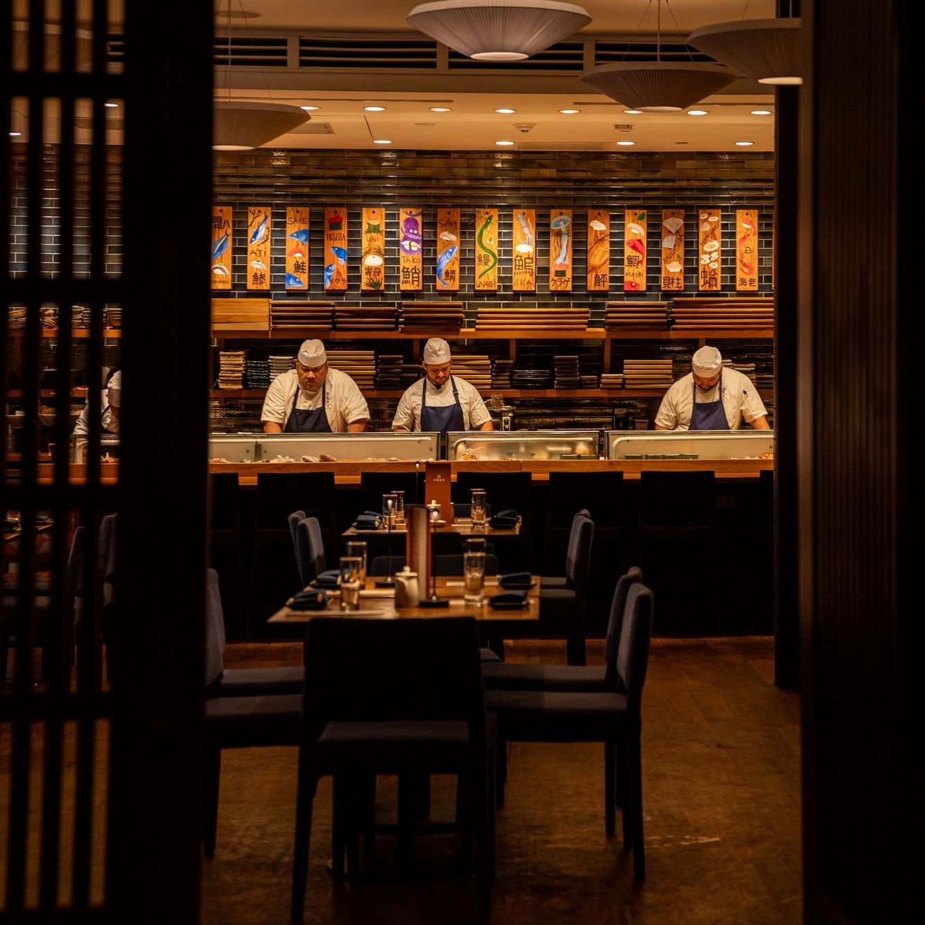 Three chefs in white uniforms and aprons prepare sushi at a modern Japanese restaurant with colorful banners and pendant lighting.