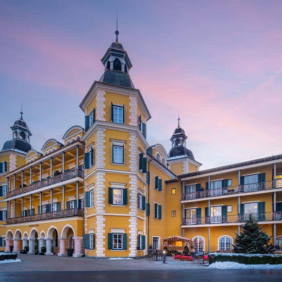 Grande edificio giallo con decorazioni e torri, circondato da neve e un albero di Natale.