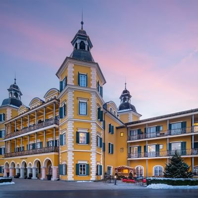 Grande edificio giallo con decorazioni e torri, circondato da neve e un albero di Natale.