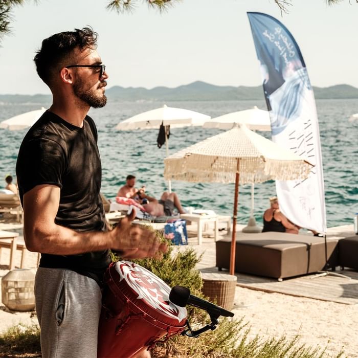 Muškarac svira crvenu bubanj pokraj plaže s ležaljkama, suncobranima i panoramskim pogledom na more.