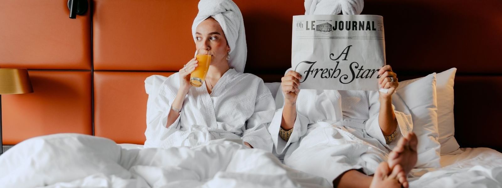 Two women in bathrobes sit on a bed, one reading a newspaper and the other drinking orange juice.