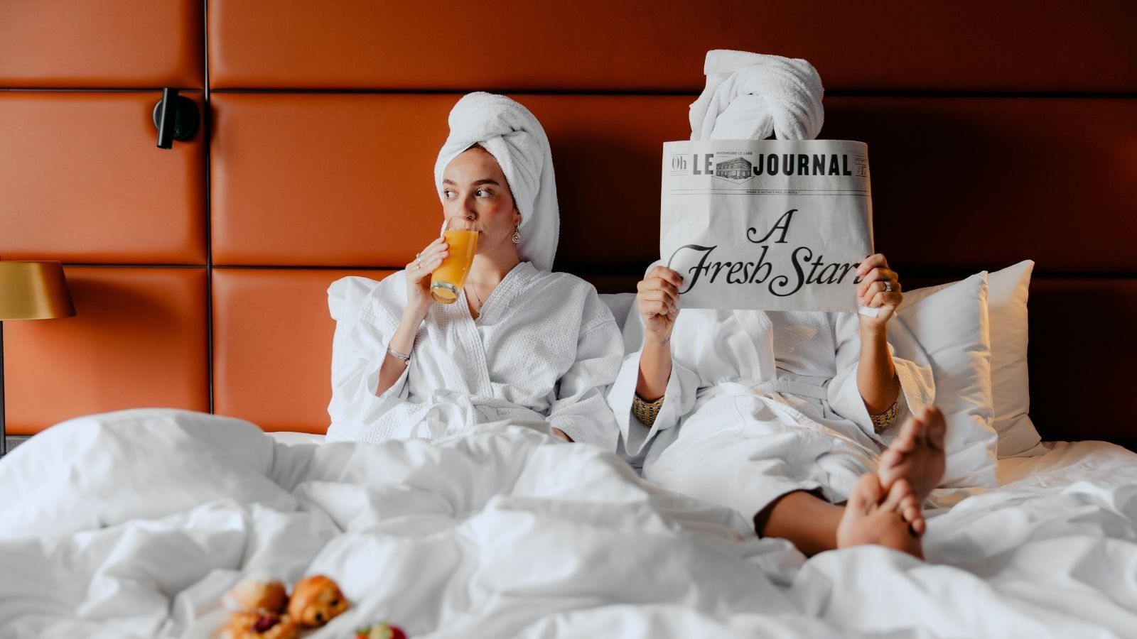 Two women in bathrobes sit on a bed, one reading a newspaper and the other drinking orange juice.