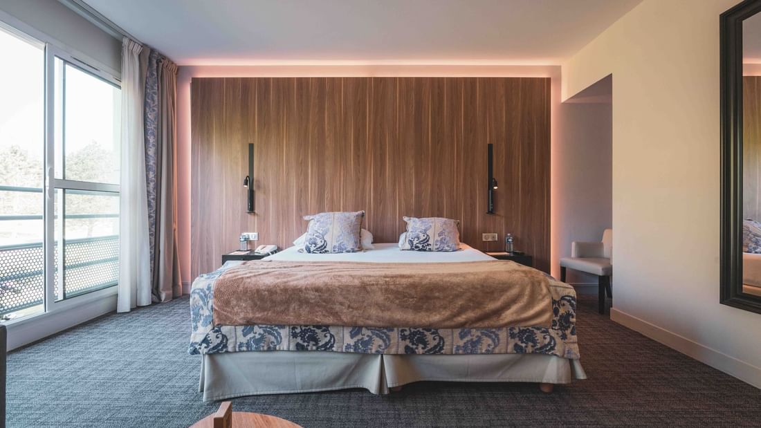 Chambre d'hôtel avec lit king-size, grands rideaux, mur en bois et vue sur l'extérieur.