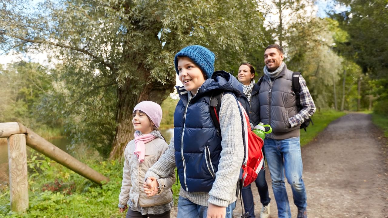 Family of four with backpacks on a forest trail, smiling and holding hands.