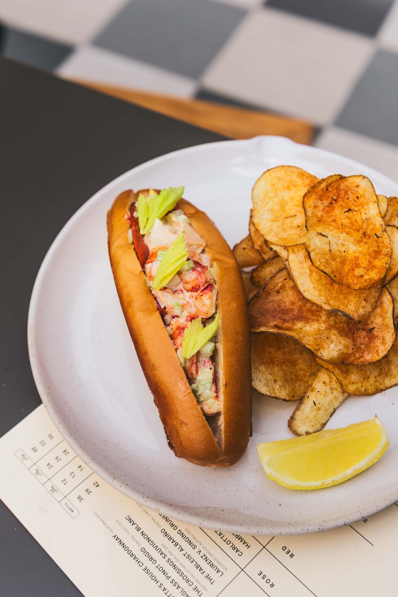 A lobster roll with lettuce on a toasted bun, served with potato chips and a lemon wedge on a white plate.