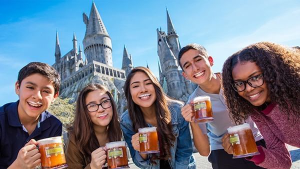 A group of five friends pose with butterbeers and white mustaches in front of Hogwarts Castle.