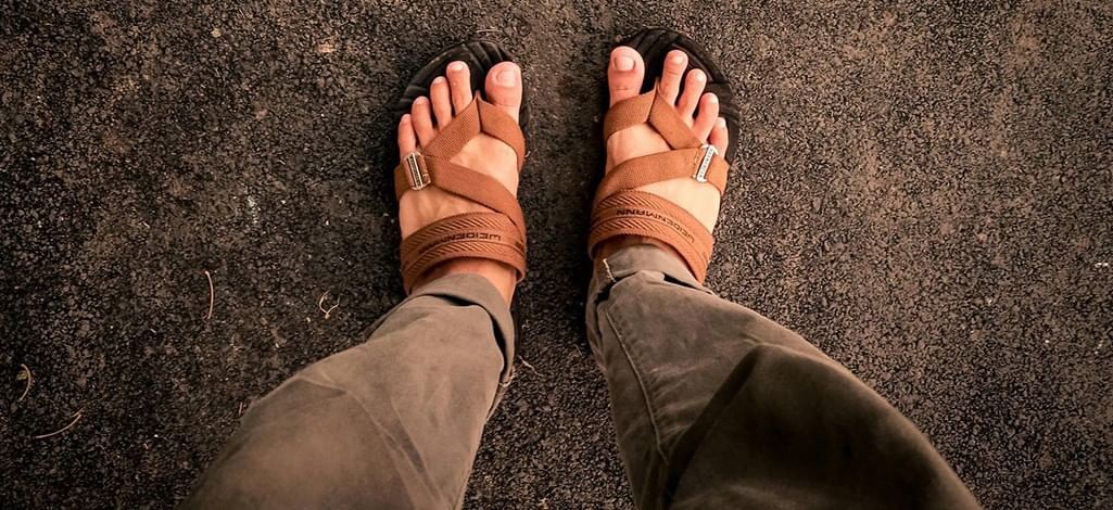 Close-up of feet wearing brown hiking sandals and khaki pants, standing on a rough dark ground.