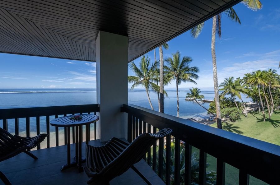 Wooden chairs by a table under a dark ceiling on a balcony with palm trees in Warwick Suites at Warwick Fiji Resort and Spa