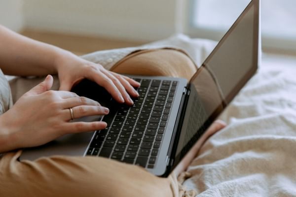 A close-up of someone sitting cross-legged on a bed with a laptop in their lap. 