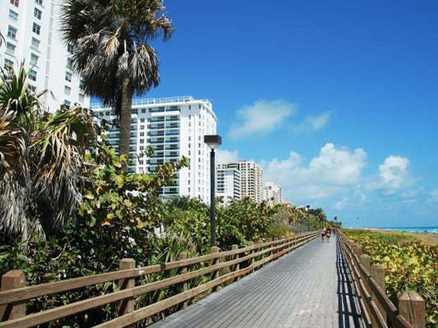 Miami Beach Boardwalk lined with a wooden fence palm trees and white high-rise buildings near Tradewinds Apartment Hotel