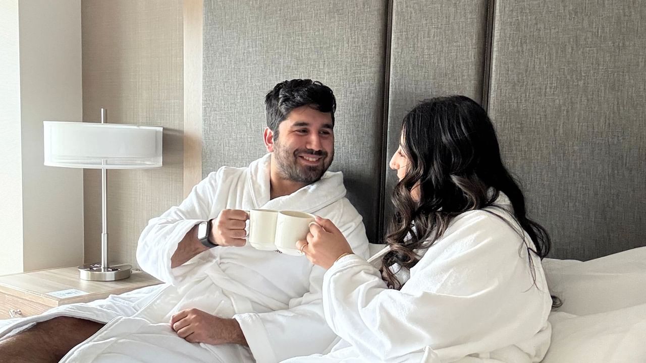 A couple in Coast Hotels bathrobes sits on a bed smiling and looking at each other.