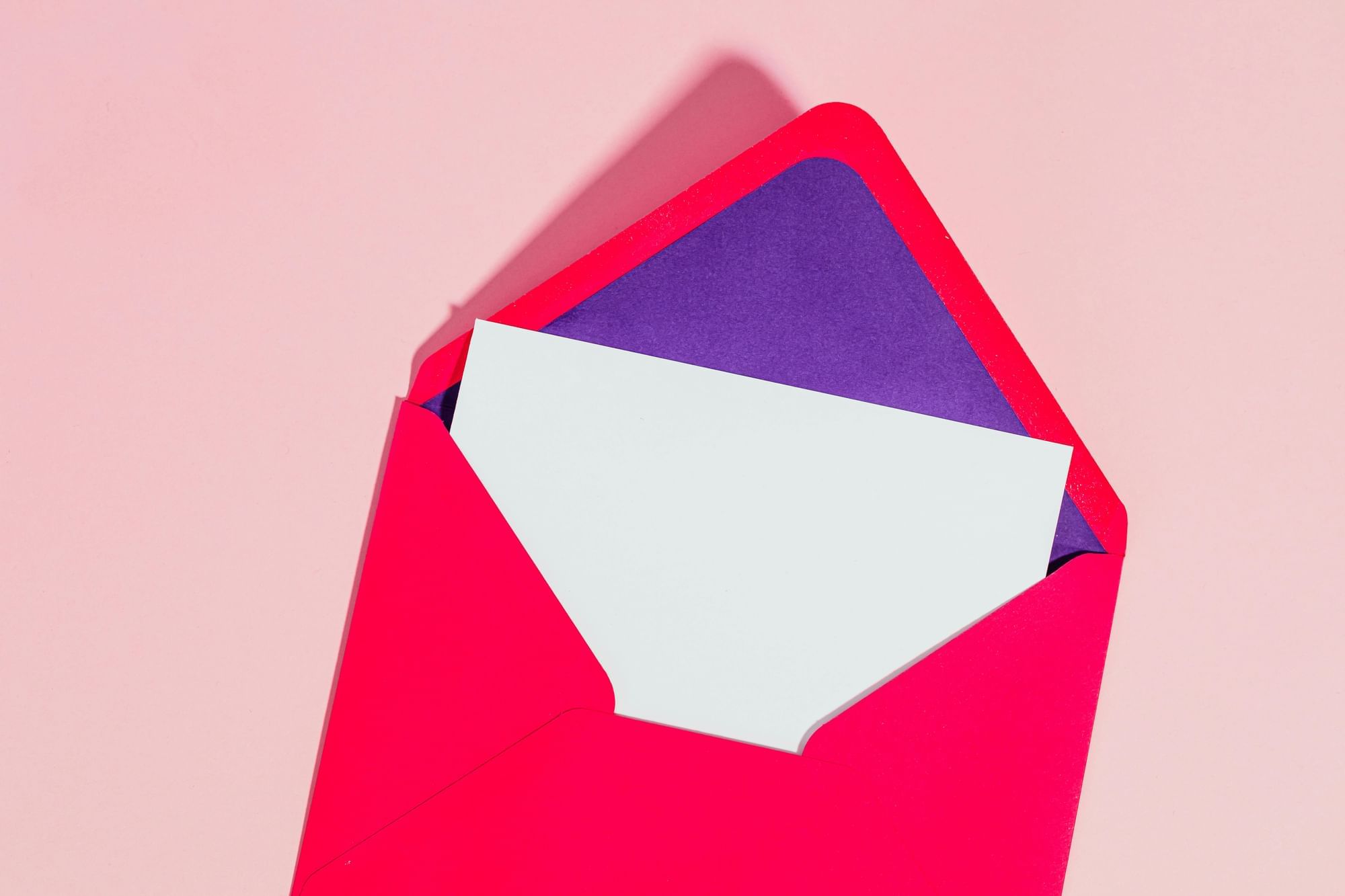 A bright pink envelope with purple lining on a pink background behind a white blank card.