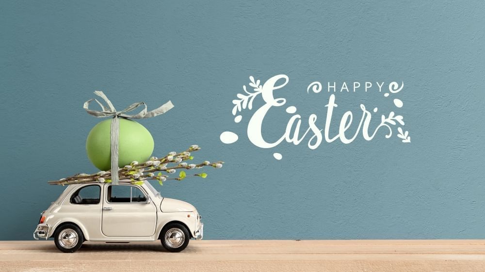 A small white toy vintage car with a bundle of willow sticks and a single green easter egg on the roof, tied with a bow against a blue background with the words Happy Easter.