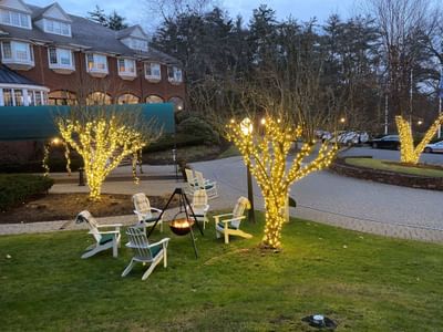 Evening lawn patio with white chairs around a cozy fire pit beside trees with fairy lights at The Simsbury Inn