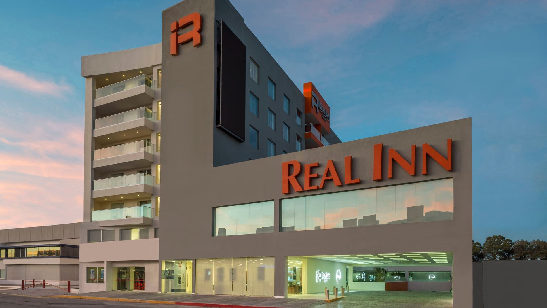 Modern facade of Real Inn Celaya at dusk, with glowing windows and the hotel name in bright orange neon