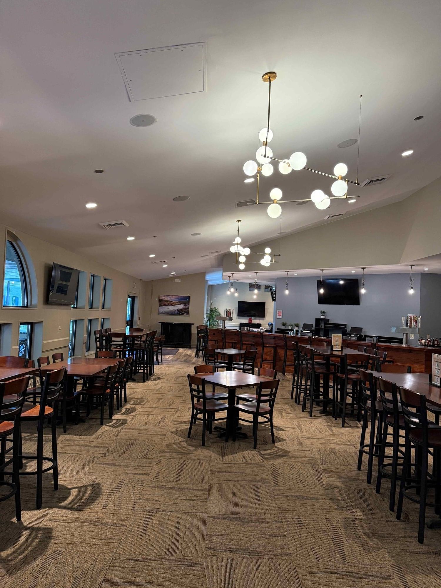 A dining area with tables, chairs, a bar, and large windows, illuminated by ceiling lights and chandeliers.