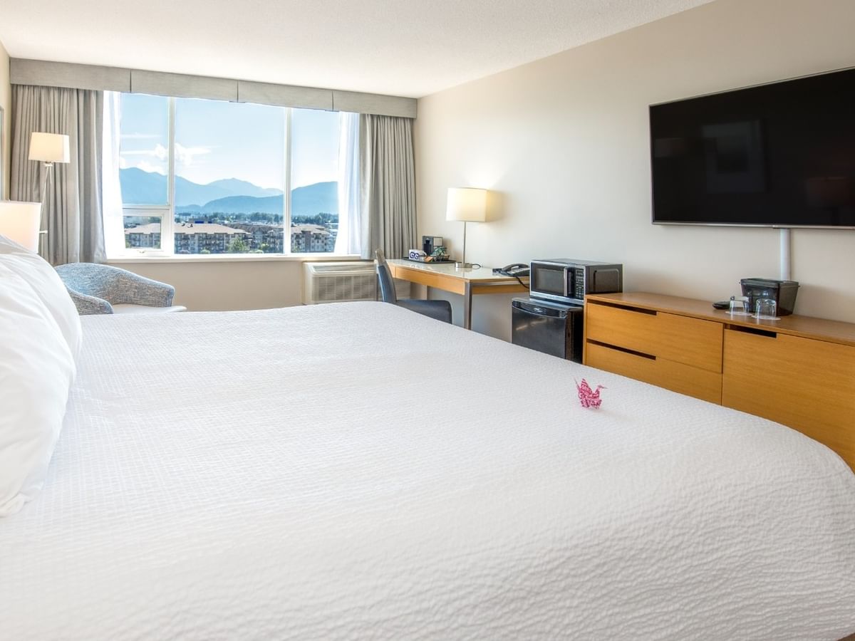 A hotel room with a large king bed, desk, TV, and window showing mountains.