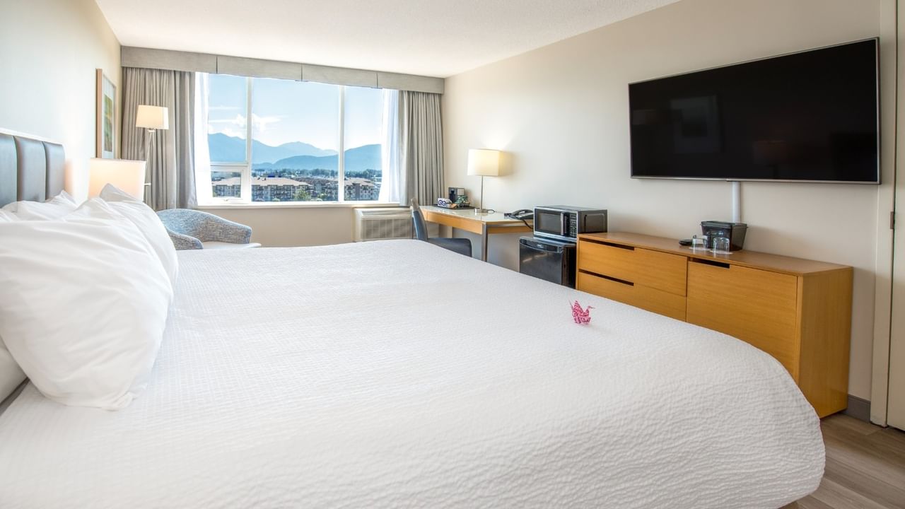 A hotel room with a large king bed, desk, TV, and window showing mountains.