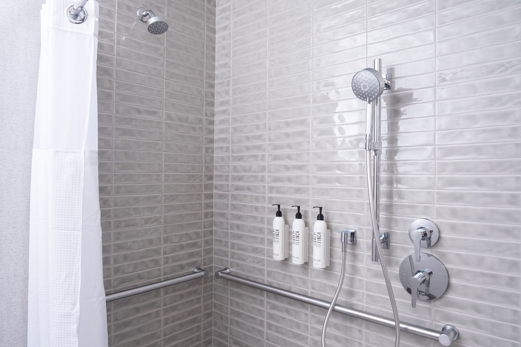 A shower with a white curtain, gray tile, and toiletries