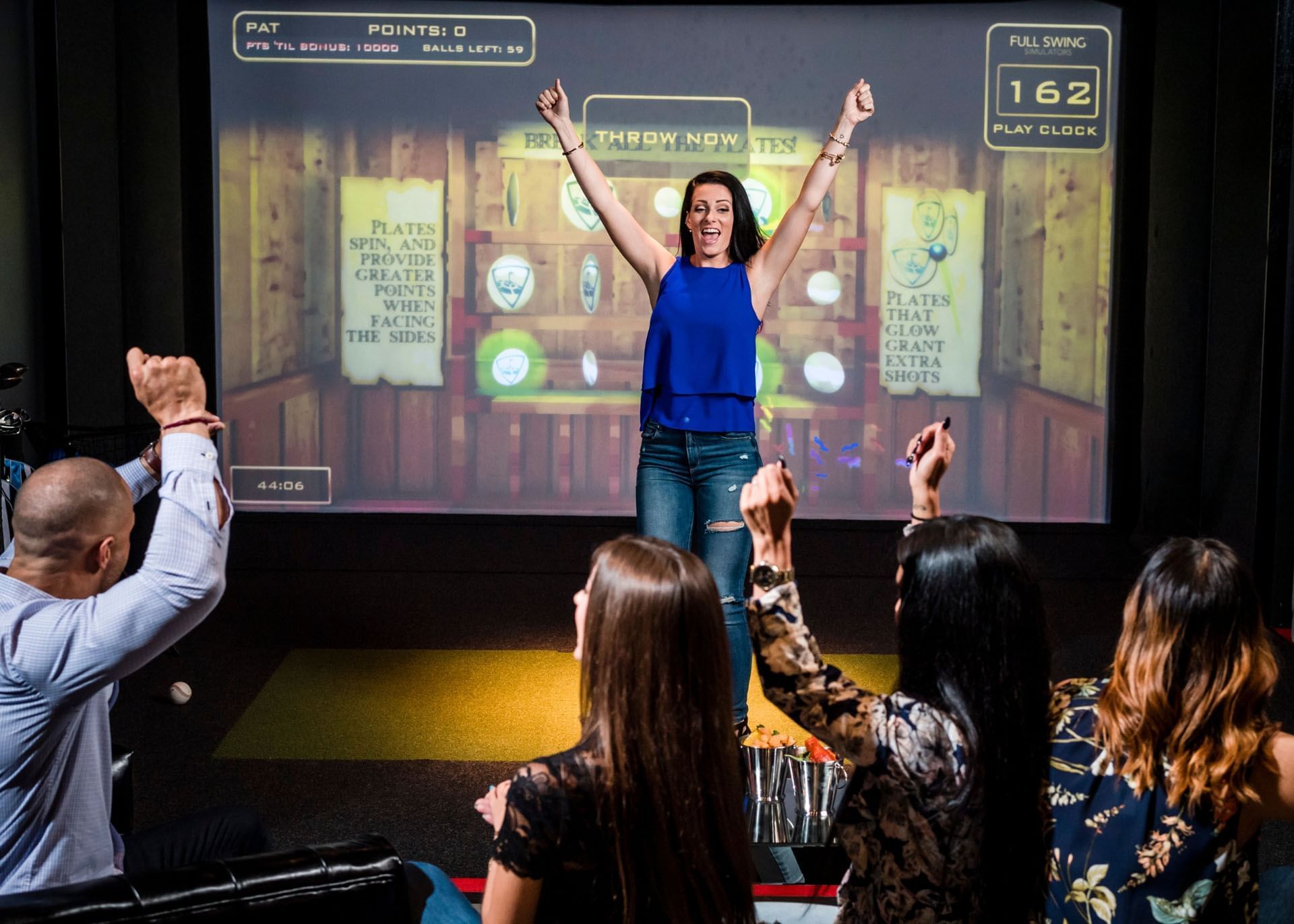 Woman celebrating by a Full Swing simulator screen, with friends cheering near cocktails at the Hotel X Toronto