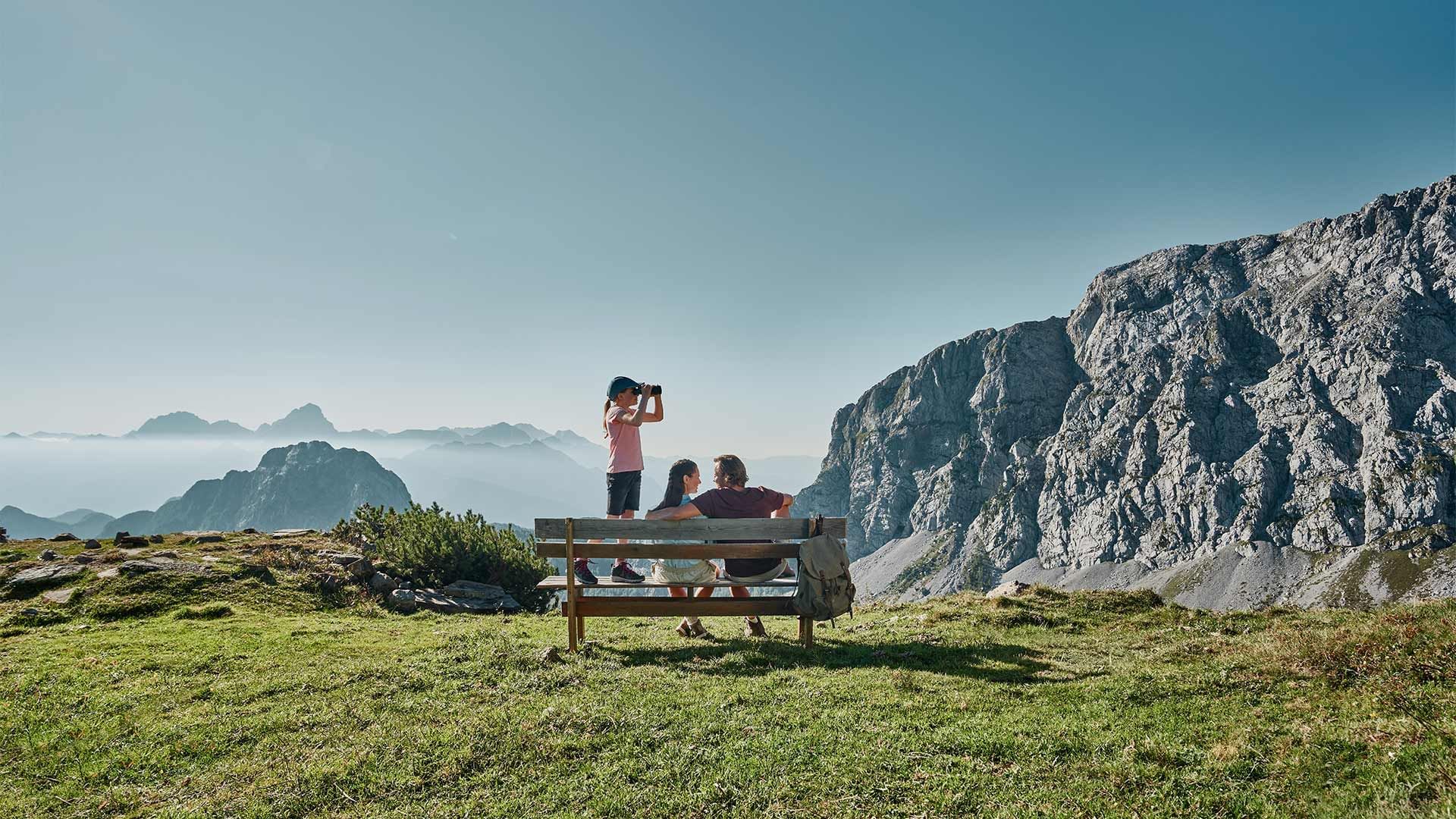 Una famiglia siede su una panchina in montagna, con un bambino che guarda attraverso un binocolo.