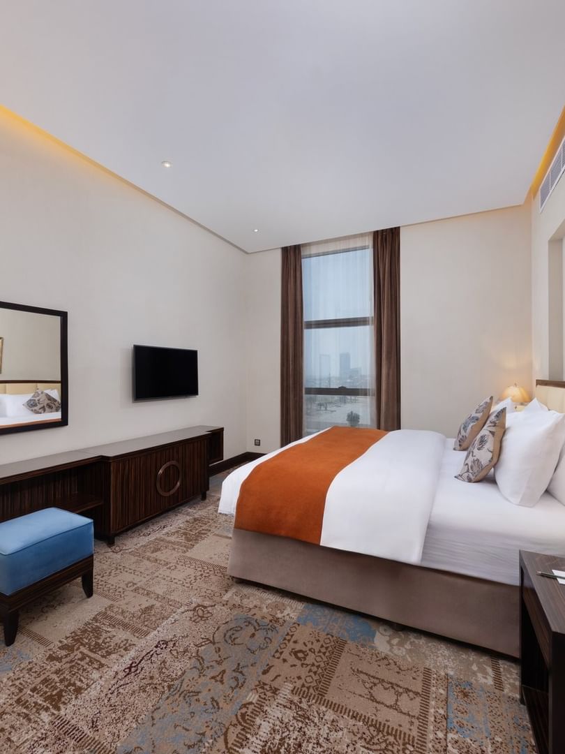 Orange bed throw by a blue velvet ottoman on a patterned rug under a wall-mounted TV in Executive Room at Warwick Al Khobar