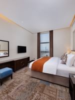 Orange bed throw by a blue velvet ottoman on a patterned rug under a wall-mounted TV in Executive Room at Warwick Al Khobar